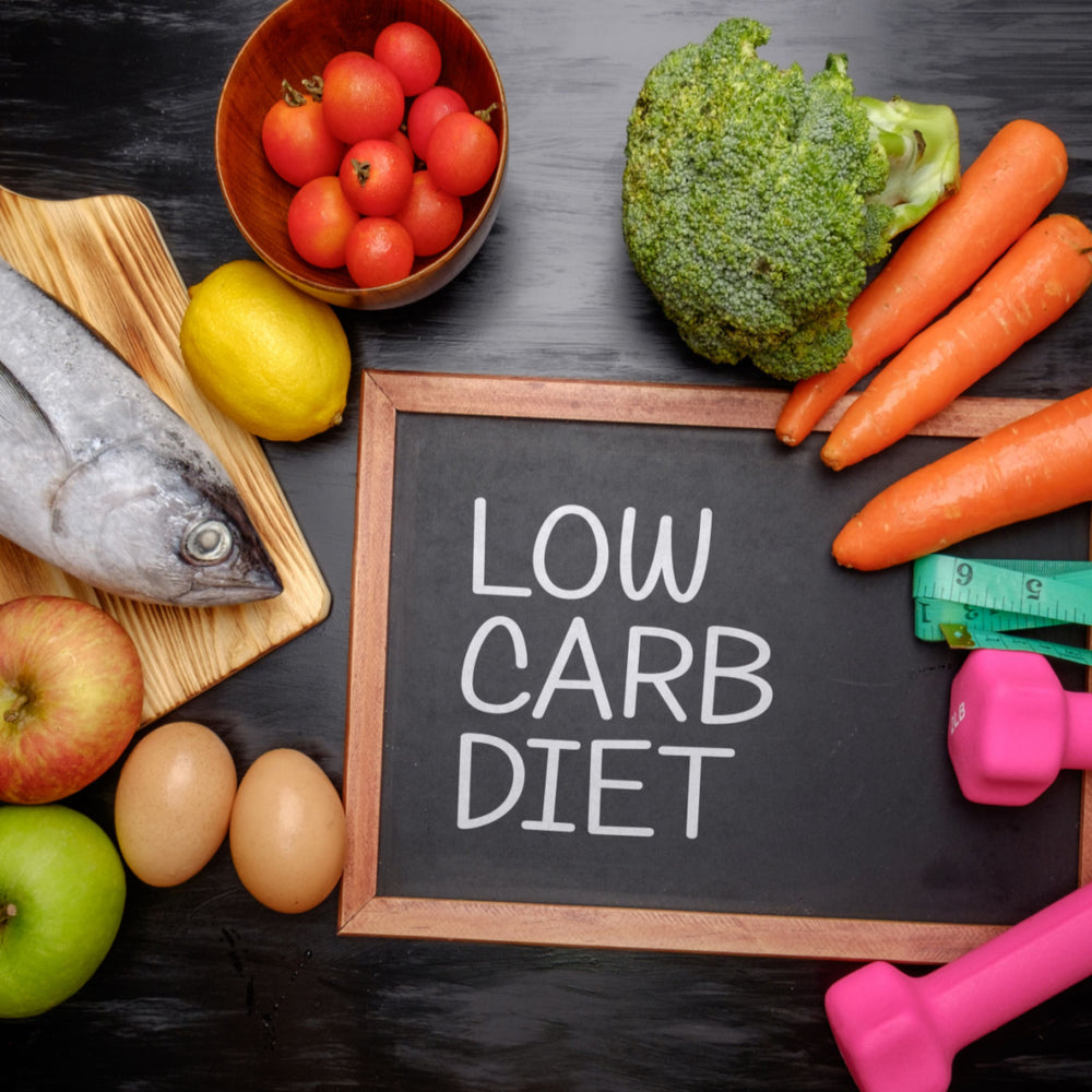 A variety of fresh foods including fish, vegetables, and fruits surround a chalkboard displaying the words "LOW CARB DIET," alongside pink dumbbells and a measuring tape.