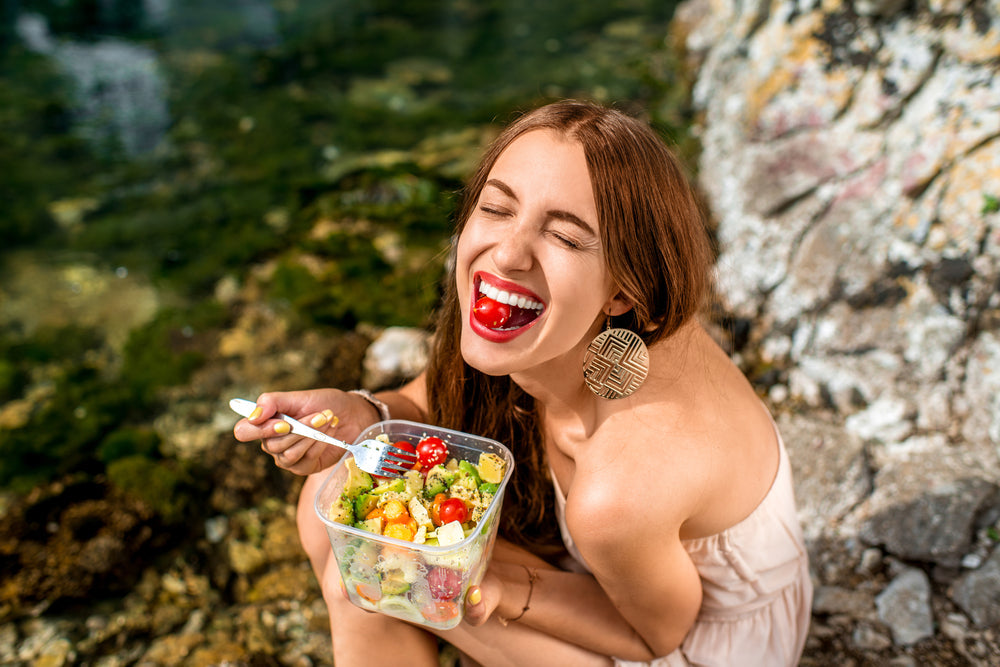 A woman with long brown hair and large earrings smiles widely while sitting on a rocky shore, holding a container of colorful salad in one hand and a cherry tomato in her mouth.