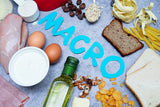 A variety of foods, including chicken, eggs, pasta, and nuts, are arranged around the word "MACRO" in blue letters on a gray surface, highlighting their role in muscle building and recovery.