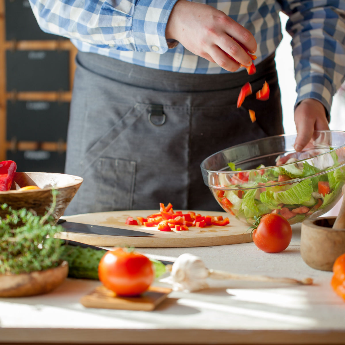 A person in a checked shirt adds chopped red peppers to a large bowl of salad greens while preparing ingredients on a kitchen counter filled with fresh vegetables and herbs.