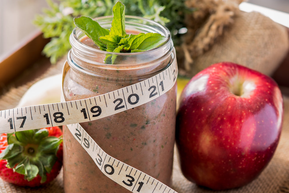 A jar of smoothie topped with mint leaves is surrounded by a measuring tape, a red apple, and a strawberry, all set on a textured surface with greenery in the background.