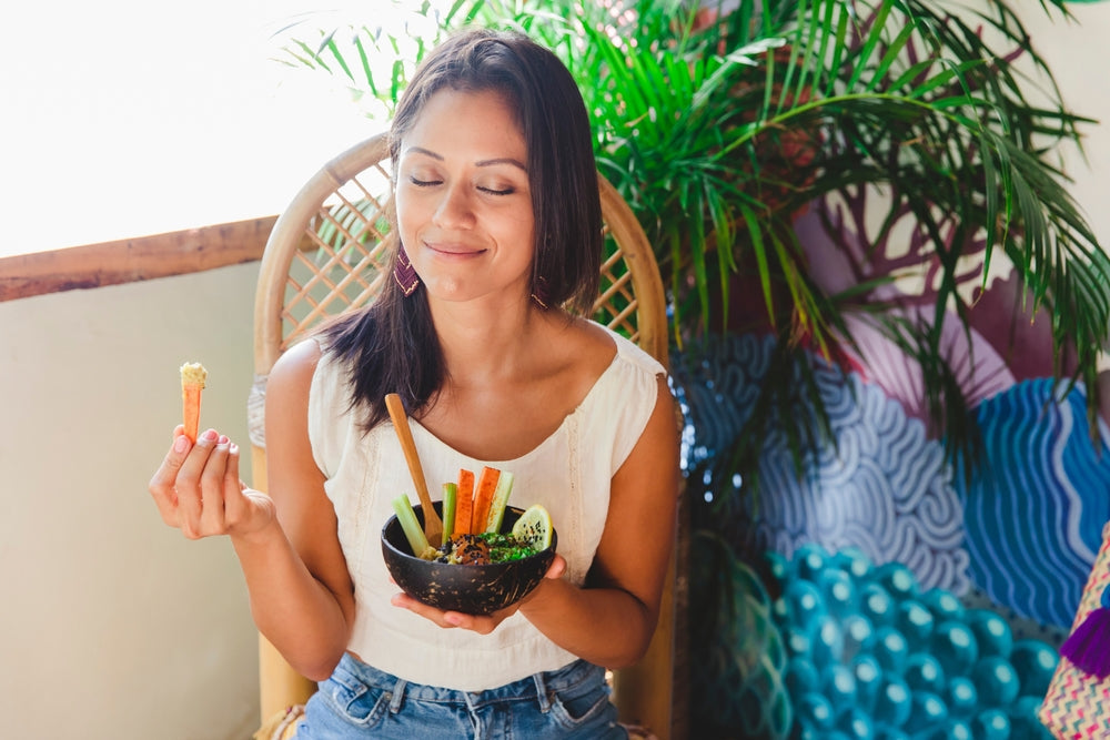 A woman with long hair sits in a rattan chair, smiling with her eyes closed while holding a carrot stick and a bowl of colorful vegetables, surrounded by indoor plants and a vibrant mural.