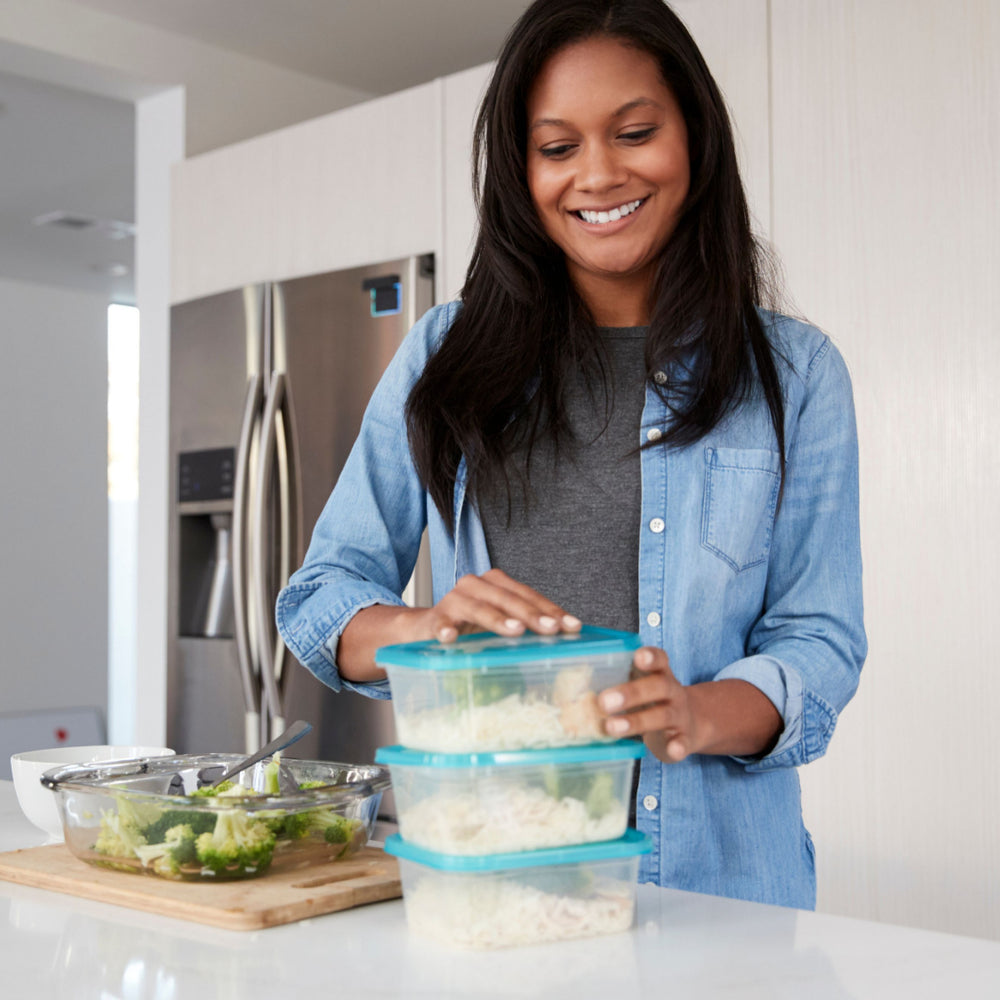 A woman with long hair smiles while placing a lid on a stack of food storage containers filled with rice in a modern kitchen, with a bowl of salad and utensils nearby.