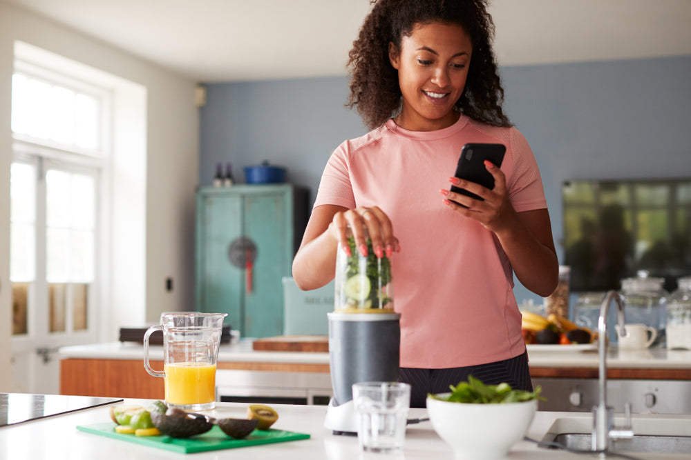A woman in a pink shirt smiles while holding a phone and preparing a smoothie with greens and fruits in a modern kitchen, featuring a pitcher of juice and a bowl of spinach nearby.