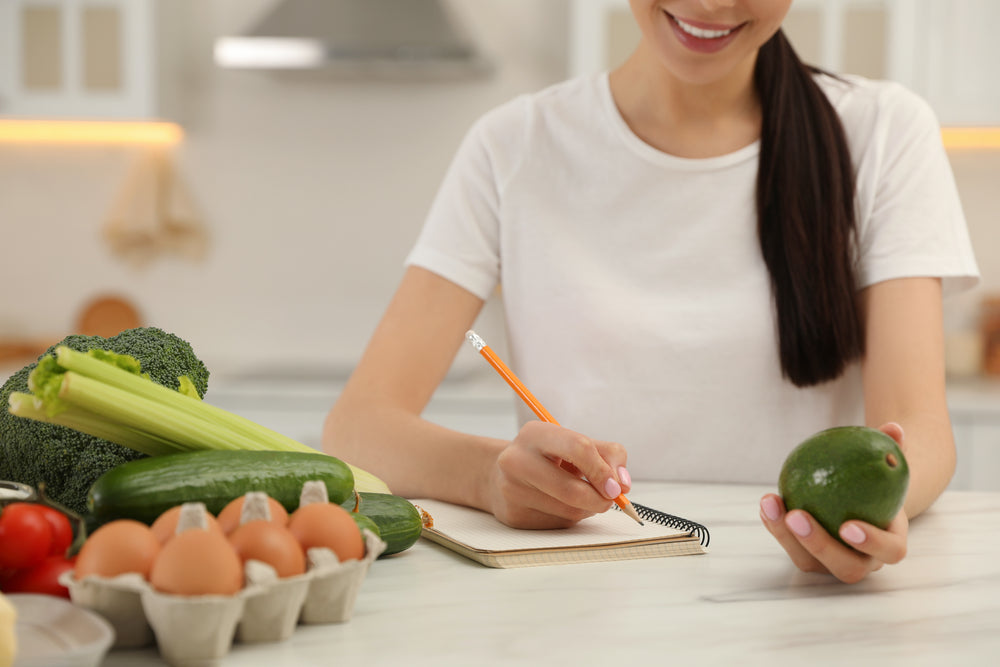 A woman in a white shirt smiles while holding an avocado in one hand and writing notes in a notebook with a pencil, surrounded by fresh vegetables and eggs on a kitchen counter.