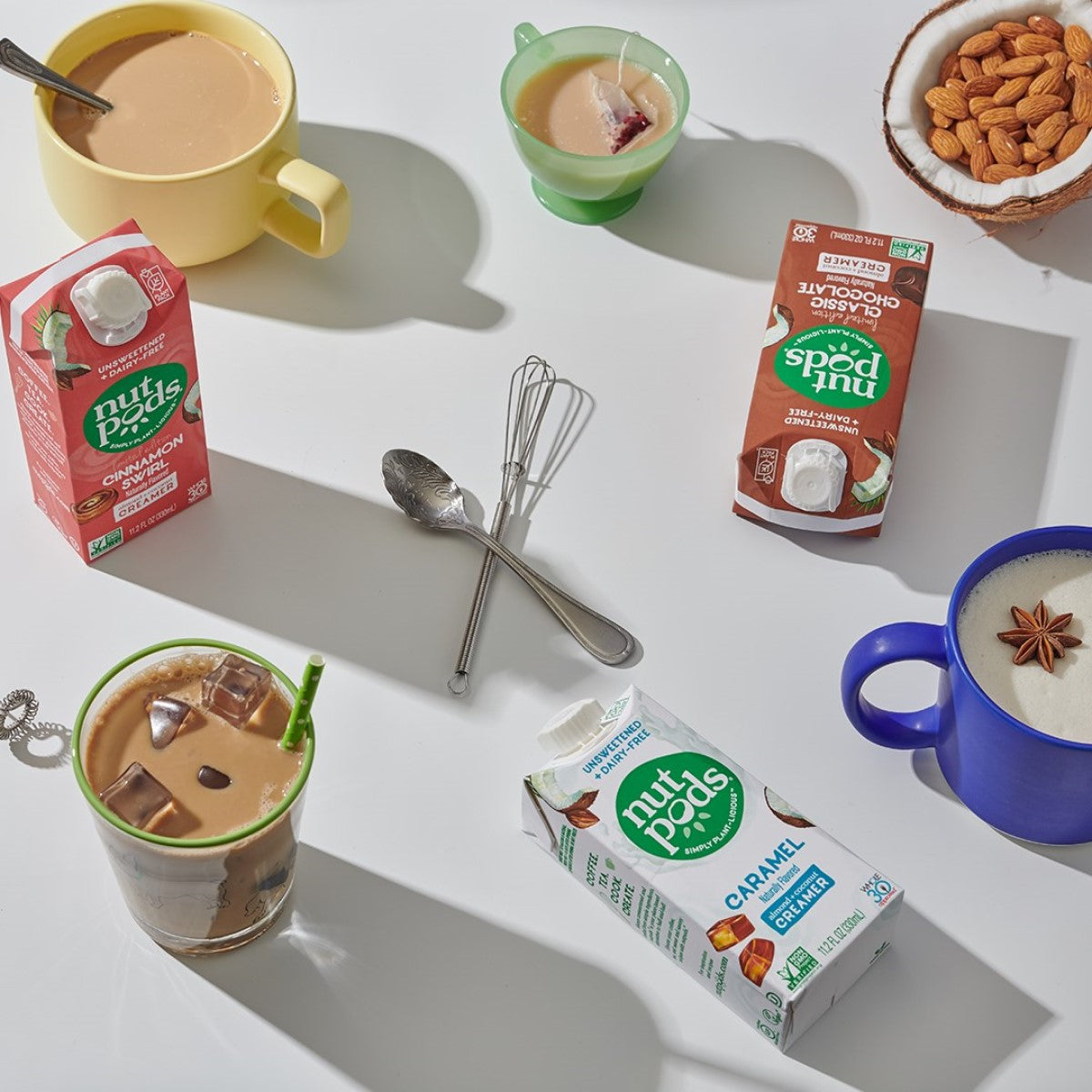 A variety of coffee drinks, including iced coffee with a green straw and mugs of hot coffee, are arranged on a white surface alongside different flavors of sugar-free creamers and a bowl of almonds.