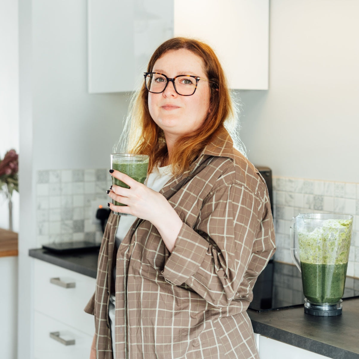 A woman with wavy red hair and glasses stands in a bright kitchen, holding a glass of green smoothie, with a blender filled with more smoothie on the counter beside her.