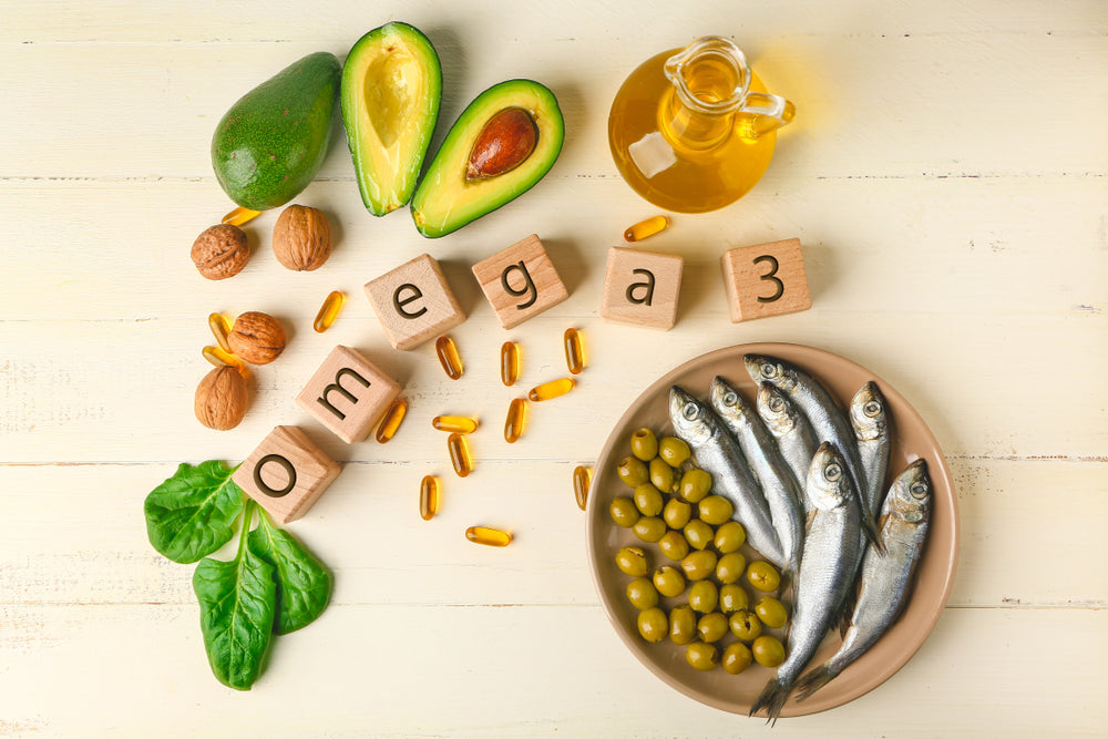 A plate of small fish and green olives is surrounded by avocados, walnuts, spinach, fish oil capsules, and wooden blocks spelling "omega 3" on a light wooden surface.