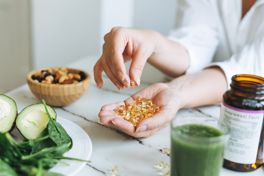 A person holds golden omega-3 capsules in one hand while reaching for more with the other, surrounded by fresh spinach, avocado, a bowl of mixed nuts, and a green smoothie on a marble table.