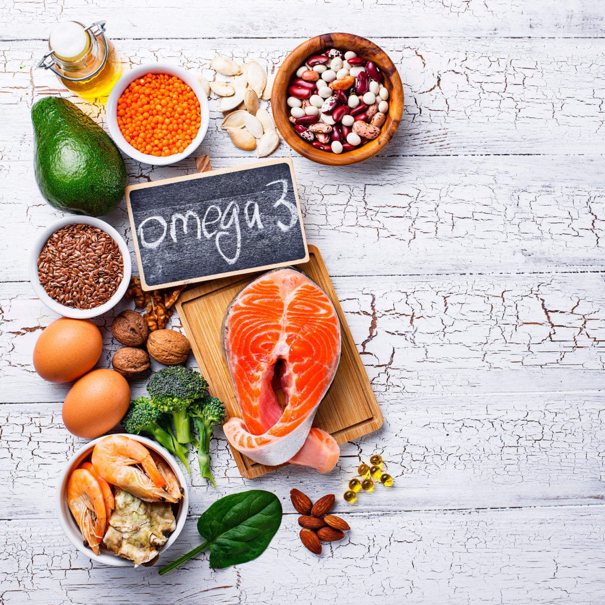 A variety of foods rich in omega-3s, including salmon, avocado, eggs, nuts, and vegetables, are arranged on a wooden surface with a chalkboard labeled "omega 3."