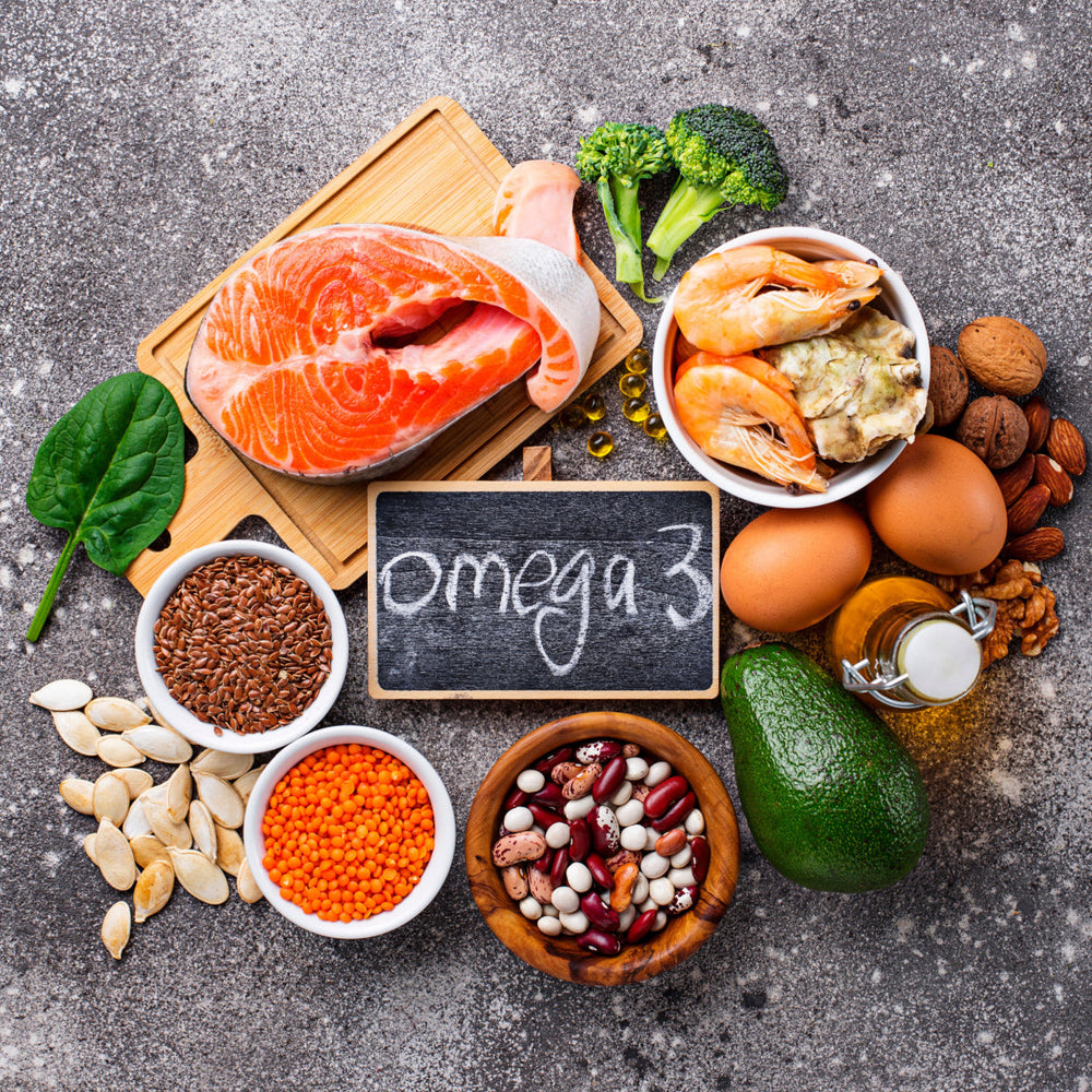 A variety of foods rich in omega-3 fats, including salmon, shrimp, eggs, nuts, seeds, and vegetables, are arranged around a chalkboard labeled "omega 3" on a textured surface.