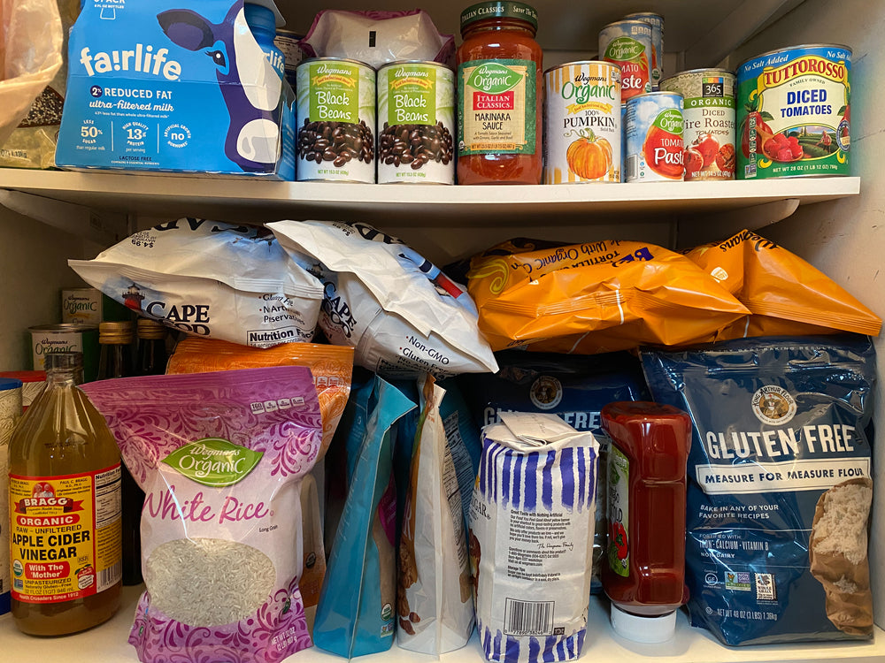 A pantry shelf is filled with various food items, including canned goods like diced tomatoes and pumpkin, bags of rice and flour, and bottles of vinegar and sauce, all organized closely together.