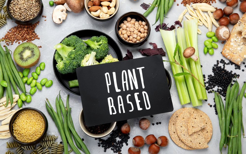 A variety of fresh vegetables, legumes, and grains surround a black sign that reads "PLANT BASED," emphasizing a healthy, plant-focused lifestyle.