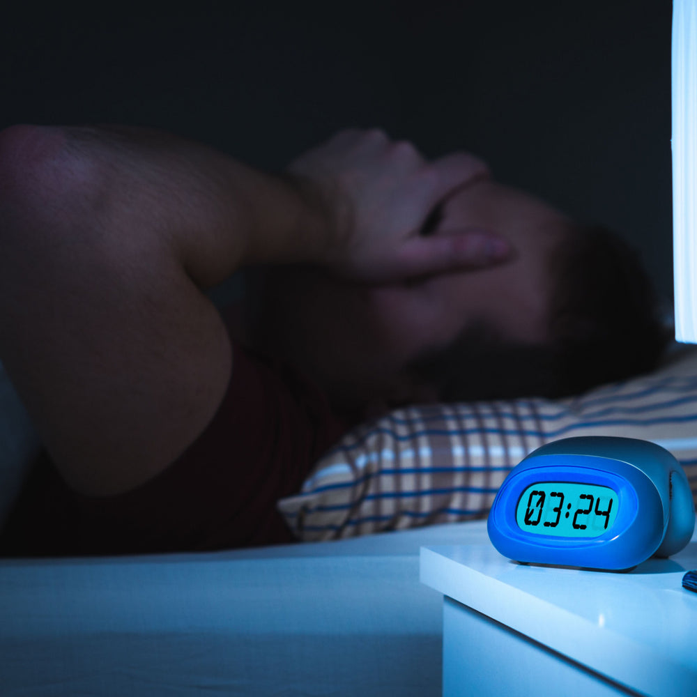 A person lies in bed with their hands covering their face, appearing distressed, while a digital clock on a nightstand shows the time as 3:24 AM in a dimly lit room.