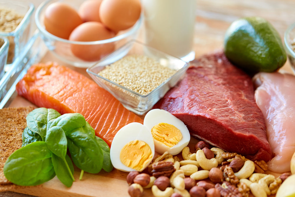 A variety of protein-rich foods, including salmon, chicken, eggs, spinach, nuts, and quinoa, are arranged on a wooden surface, highlighting their importance in a balanced diet.
