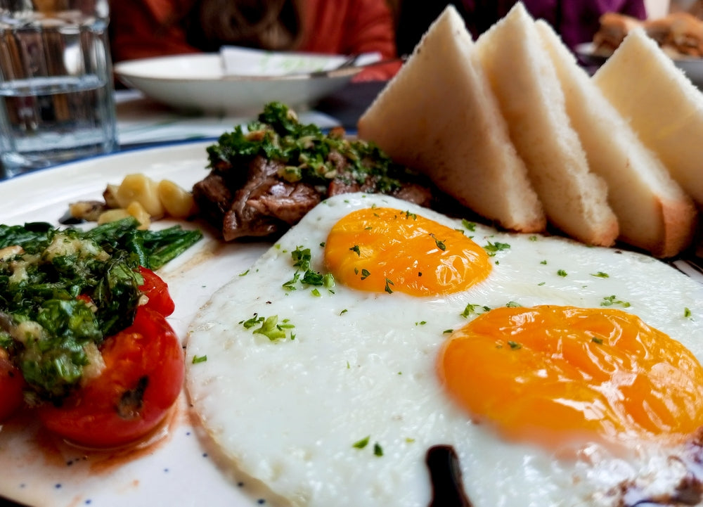 A plate features two sunny-side-up eggs with bright yellow yolks, sautéed greens and tomatoes, a piece of meat topped with herbs, and slices of white bread, set in a casual dining environment.