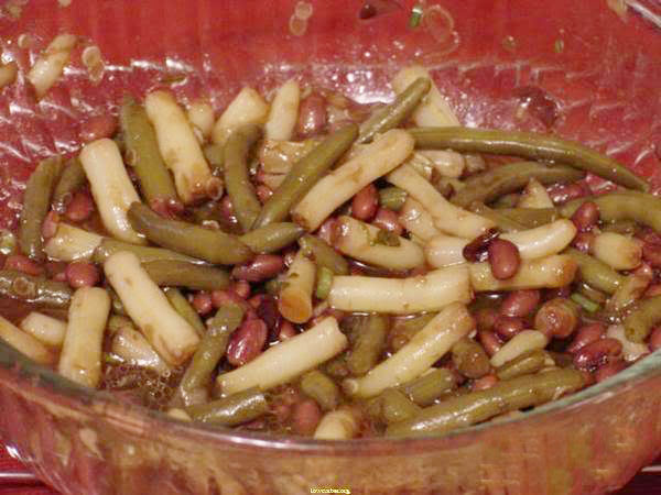 A glass bowl contains a colorful mix of green beans, wax beans, and kidney beans, tossed together in a light dressing, set against a red background.