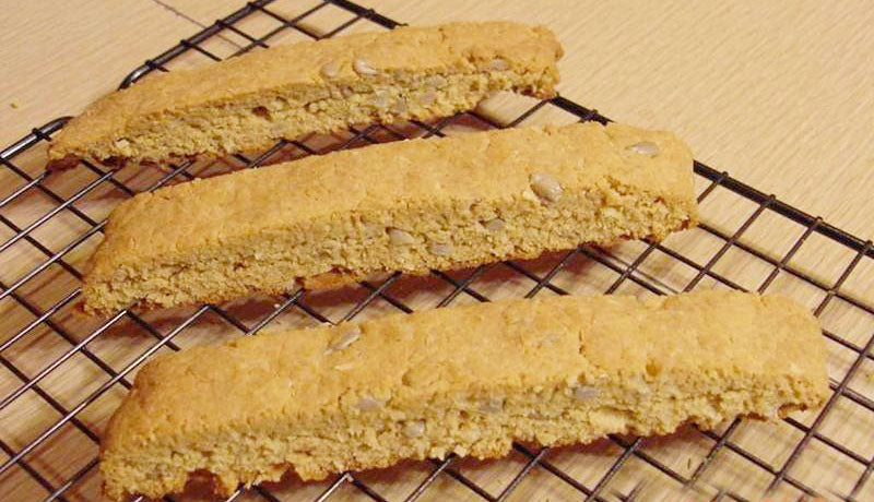 Three golden-brown biscotti with a crunchy texture are cooling on a wire rack, showcasing a light sprinkle of seeds on top against a wooden background.