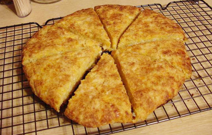 A round, golden-brown cornbread is cut into eight wedges and placed on a wire cooling rack, showcasing its crispy top and soft interior.