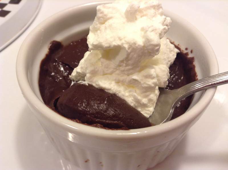 A white ramekin filled with rich chocolate mousse topped with a generous swirl of whipped cream, with a silver spoon resting on the edge.