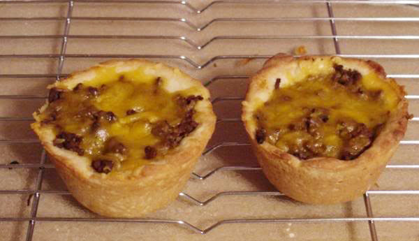 Two baked meat and cheese cups sit on a wire cooling rack, showcasing a golden crust filled with ground beef and melted cheddar cheese.