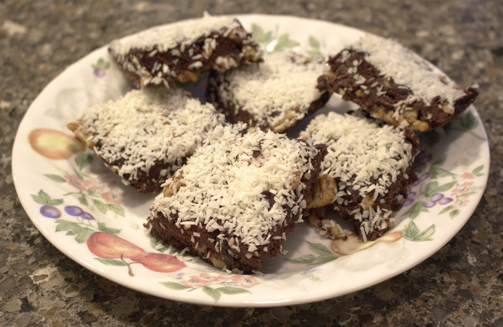 A plate of chocolate coconut bars topped with shredded coconut and walnuts is arranged on a decorative floral plate, resting on a dark stone countertop.