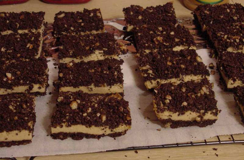 A tray of neatly cut dessert bars features layers of chocolate crumb topping and a creamy, light-colored filling, arranged on a sheet of parchment paper.