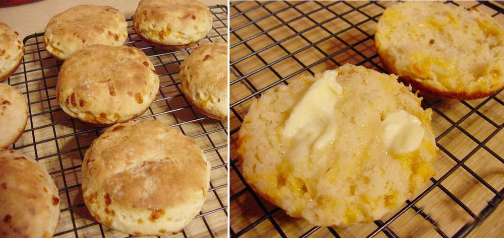 Freshly baked cheese biscuits are cooling on a wire rack, with one biscuit split open and spread with butter, showcasing a fluffy interior and golden crust.
