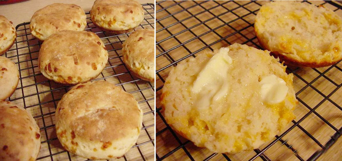 Freshly baked cheese biscuits are cooling on a wire rack, with one biscuit split open and spread with butter, showcasing a fluffy interior and golden crust.