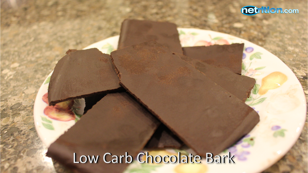A plate of low-carb chocolate bark pieces is arranged on a floral-patterned dish, showcasing their rich dark color against a textured countertop.