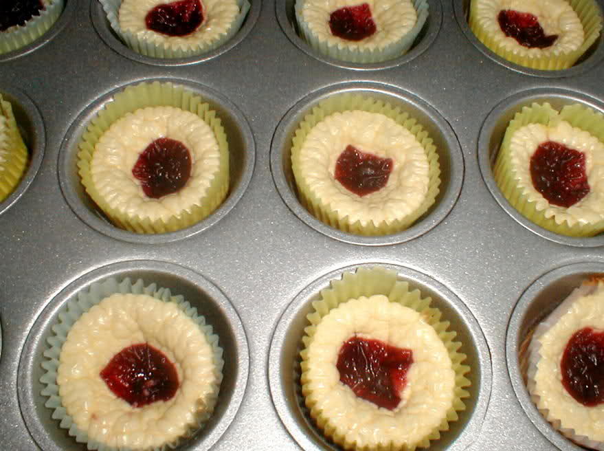 A muffin tin holds several unbaked cheesecake cups with a dollop of red fruit filling in the center, surrounded by green cupcake liners.