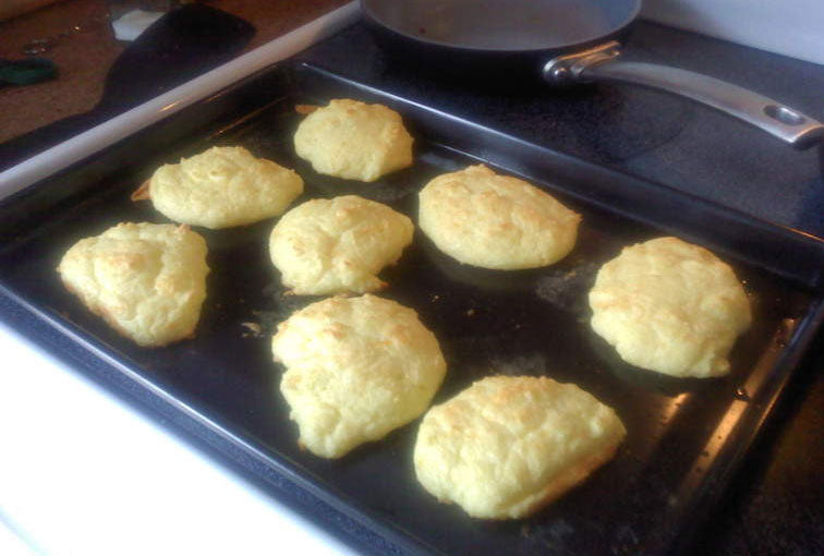 Golden-brown, fluffy cheese rolls are arranged on a black baking sheet, with a frying pan in the background, showcasing a low-carb, gluten-free option for bread.