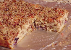 A freshly baked coffee cake with a crumbly pecan topping sits on a silver foil surface, showing a slice removed to reveal its moist interior with hints of fruit.