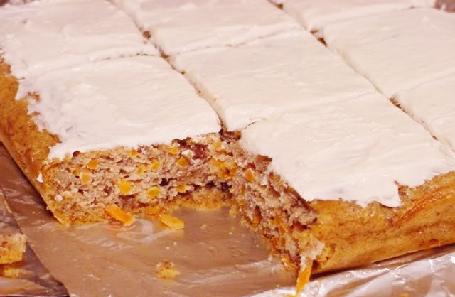 A rectangular cake with a creamy white frosting is cut into squares, revealing a moist interior with bits of orange, placed on a silver foil surface.