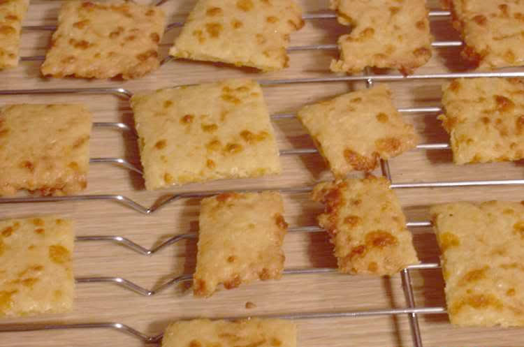 Golden-brown, square cheese crackers are cooling on a wire rack, some are partially broken, with a wooden surface visible beneath them.