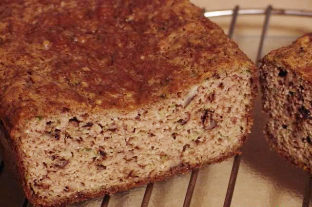 A freshly baked loaf of bread, with a golden-brown crust and a soft, textured interior, sits on a wire cooling rack, showcasing its moist crumb and flecks of herbs or ingredients.