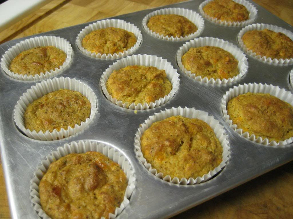 A metal muffin tin holds twelve freshly baked muffins in paper liners, with a golden-brown color and a slightly domed shape, resting on a wooden surface.