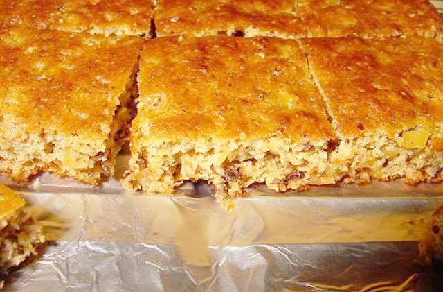 A freshly baked, golden-brown cornbread is cut into squares, revealing a moist texture with visible pieces of corn and nuts, resting on a silver tray.