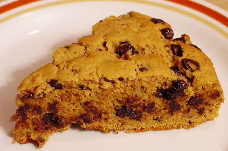A slice of chocolate chip cookie cake is displayed on a white plate with a decorative rim, showcasing its golden-brown exterior and gooey chocolate chunks throughout.