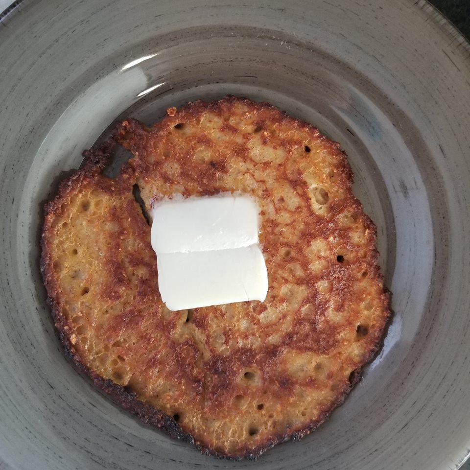 A golden-brown pancake sits in a gray bowl, topped with two squares of melting butter, indicating a warm and inviting breakfast setting.