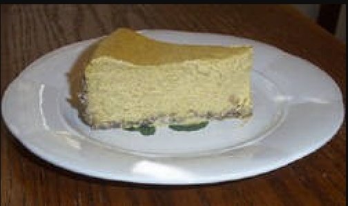A slice of creamy pumpkin cheesecake with a crumbly crust sits on a white plate, resting on a wooden table.