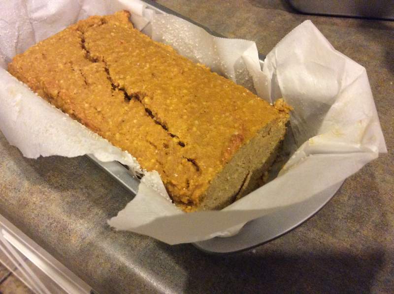 A freshly baked loaf of bread sits in a metal pan lined with parchment paper, showcasing a golden-brown top with a crack running down the center, on a textured countertop.
