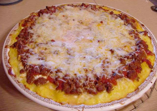 A round, cheesy pizza topped with ground meat and sauce sits on a decorative plate against a light-colored table surface.