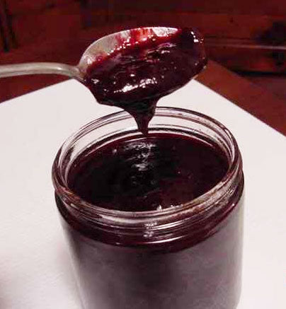 A spoon scoops out thick, dark purple jam from a glass jar, with a white surface underneath, suggesting a kitchen setting.