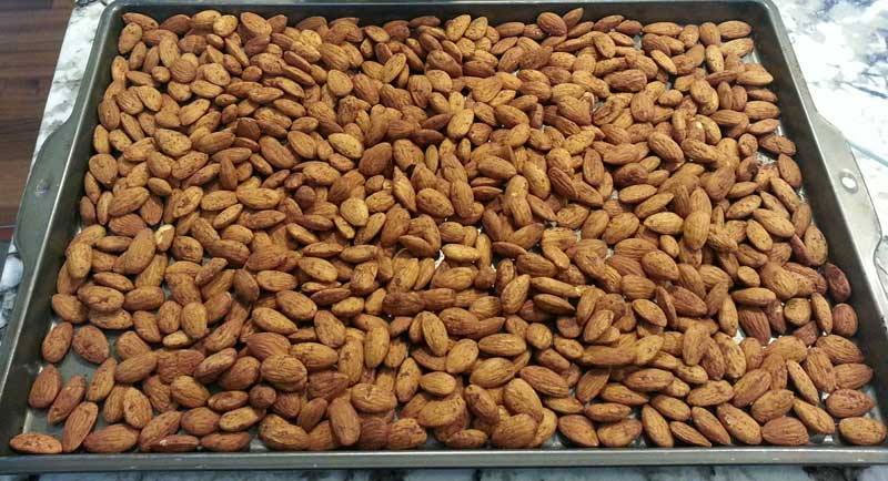 A metal baking tray is filled with a dense layer of roasted almonds, showcasing their brown, textured shells against a light-colored countertop.
