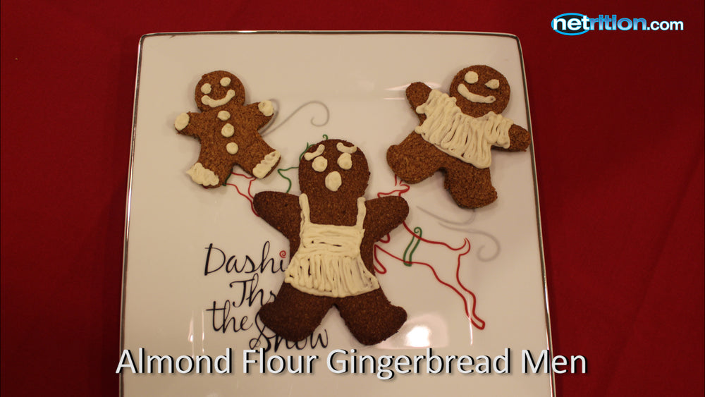 Three gingerbread men cookies, decorated with icing, are arranged on a white plate with festive designs, set against a red background, labeled "Almond Flour Gingerbread Men."