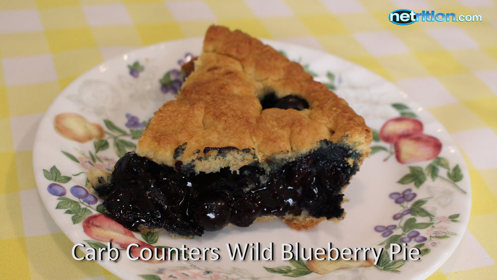 A slice of wild blueberry pie sits on a decorative plate, showcasing a golden crust and dark, glossy filling, against a yellow checkered tablecloth background.