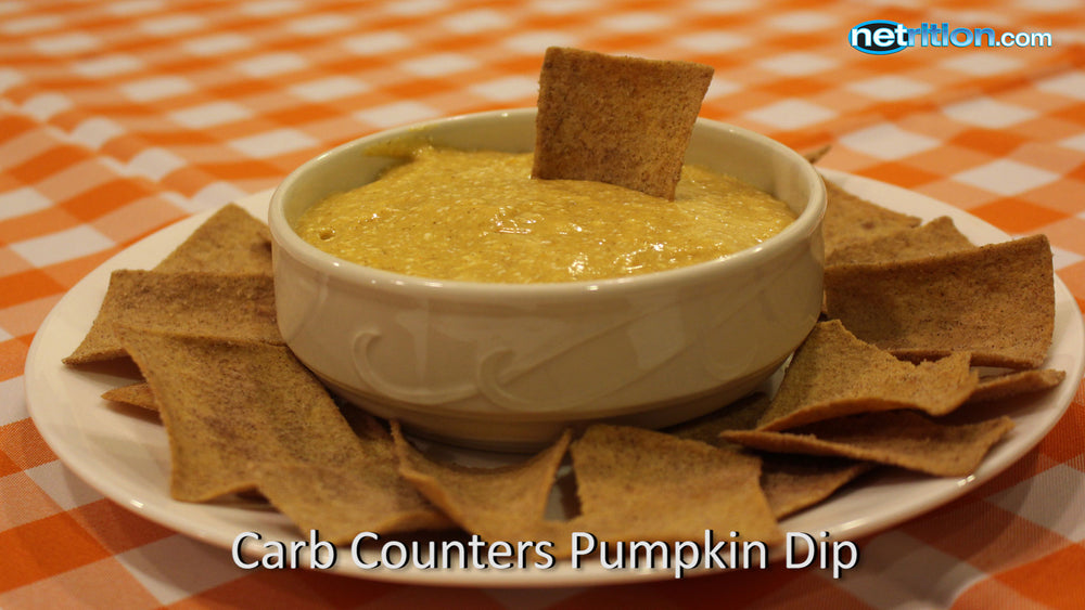 A white bowl filled with pumpkin dip sits in the center of a plate surrounded by crispy, brown chips, all on a checkered orange and white tablecloth.