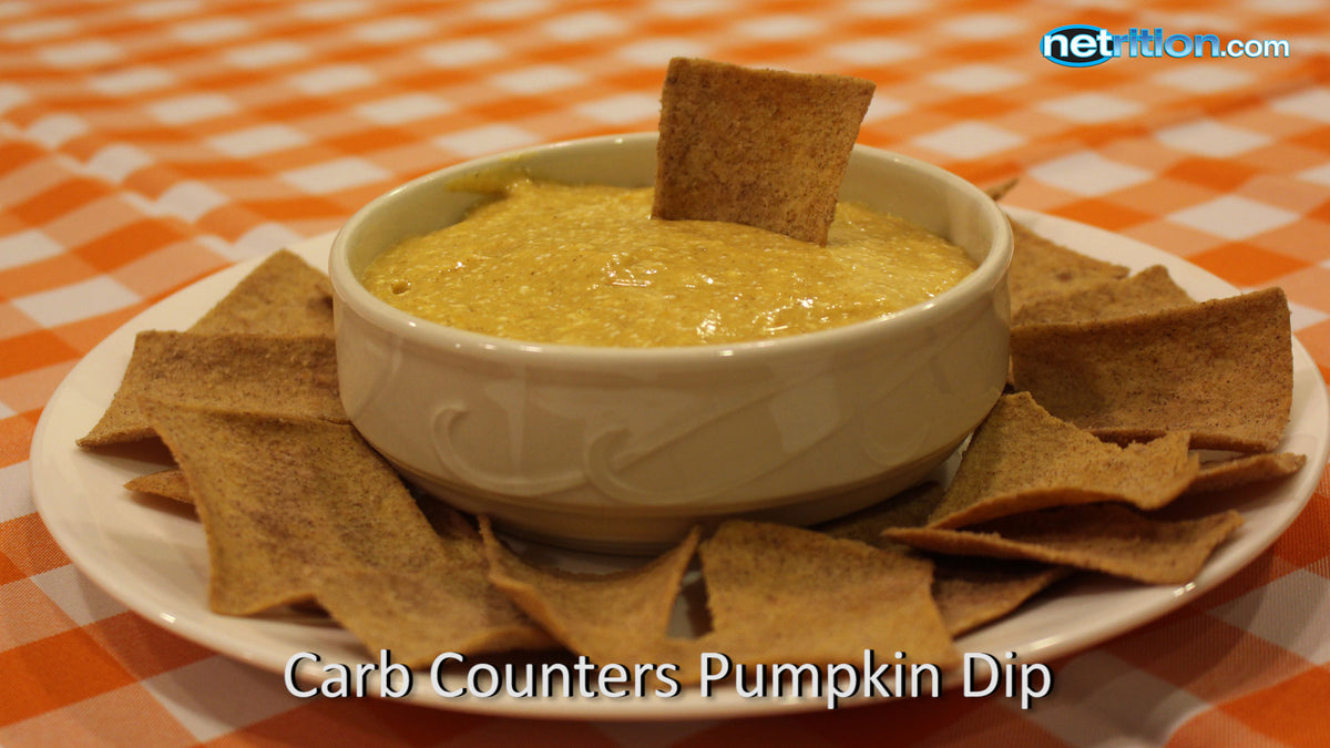 A white bowl filled with pumpkin dip sits in the center of a plate surrounded by crispy, brown chips, all on a checkered orange and white tablecloth.