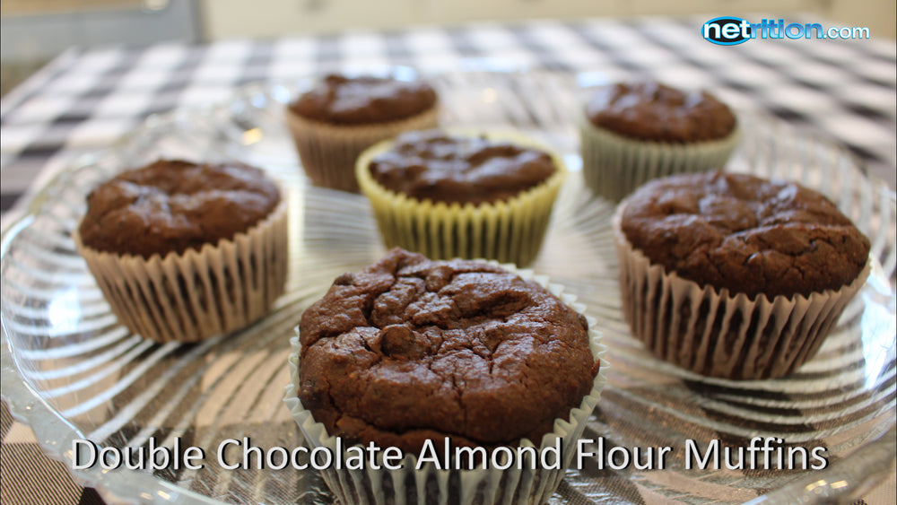 A plate of double chocolate almond flour muffins sits on a glass dish, showcasing several muffins with varying wrappers, against a checkered tablecloth background.